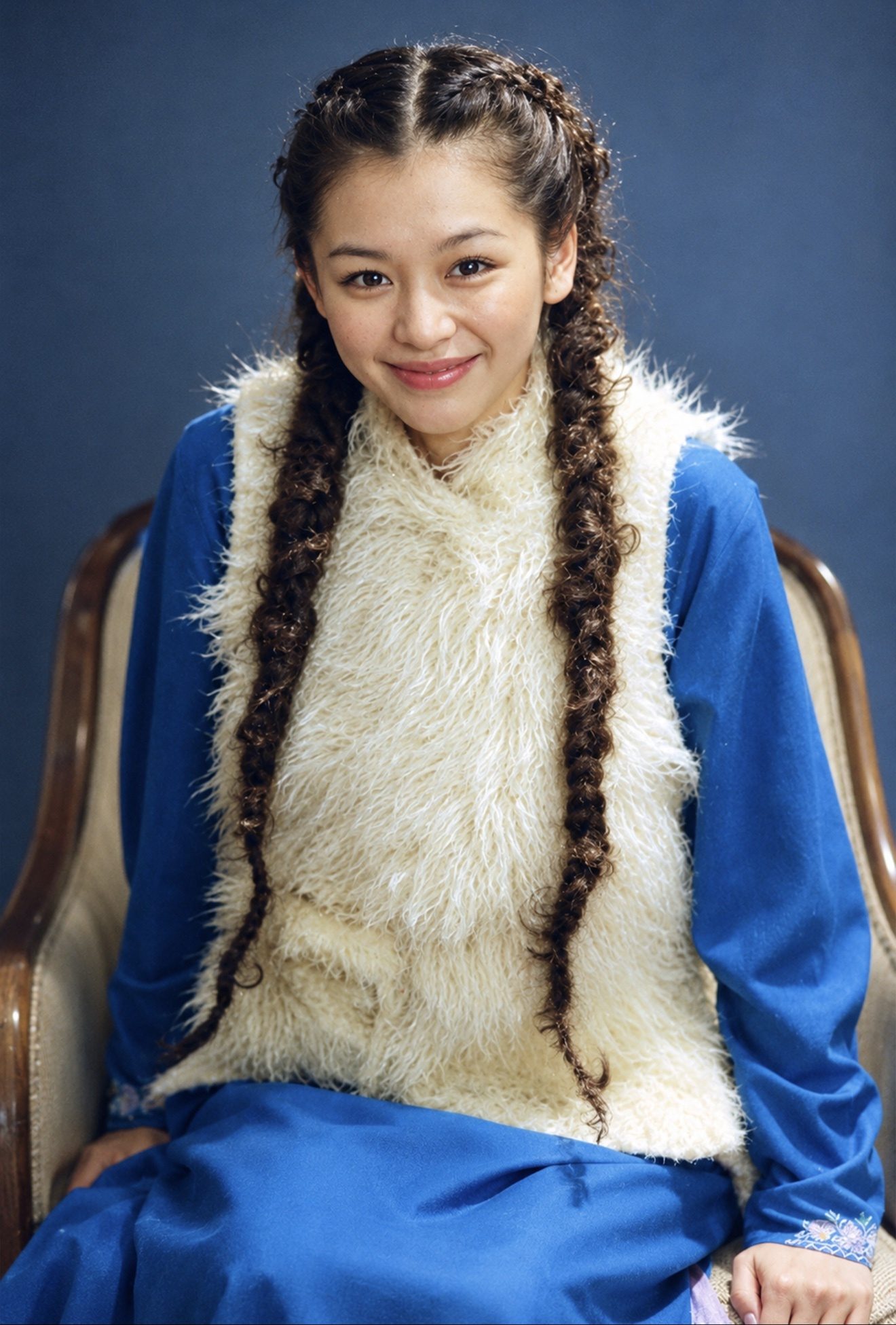 彩色照片：身穿蓝色衣衫与白色毛背心、梳着两条长辫的女子坐在椅中微笑望向镜头