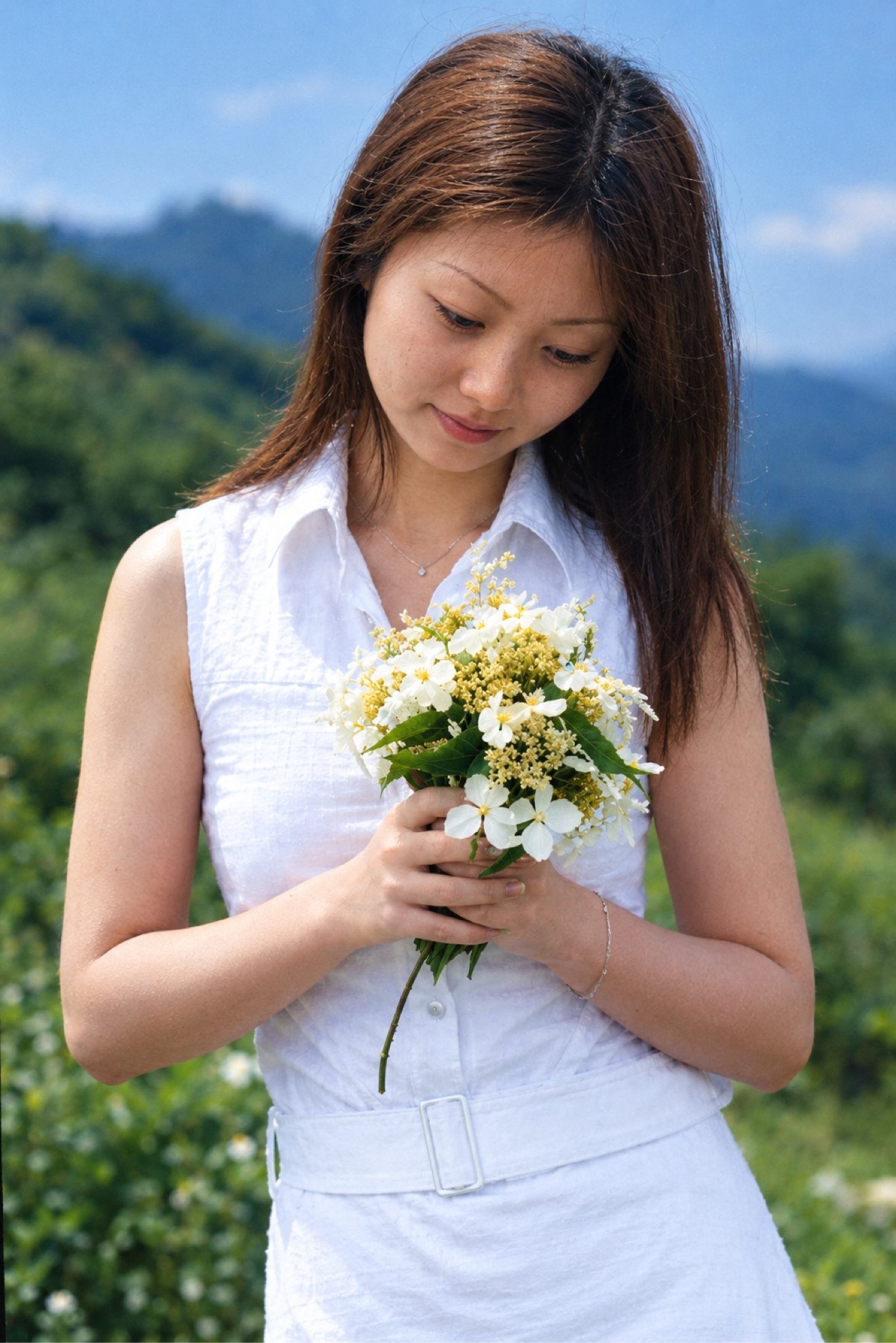 彩色照片：身穿白色无袖连衣裙的长发女子手捧一束白花，低头含笑，户外青山为背景