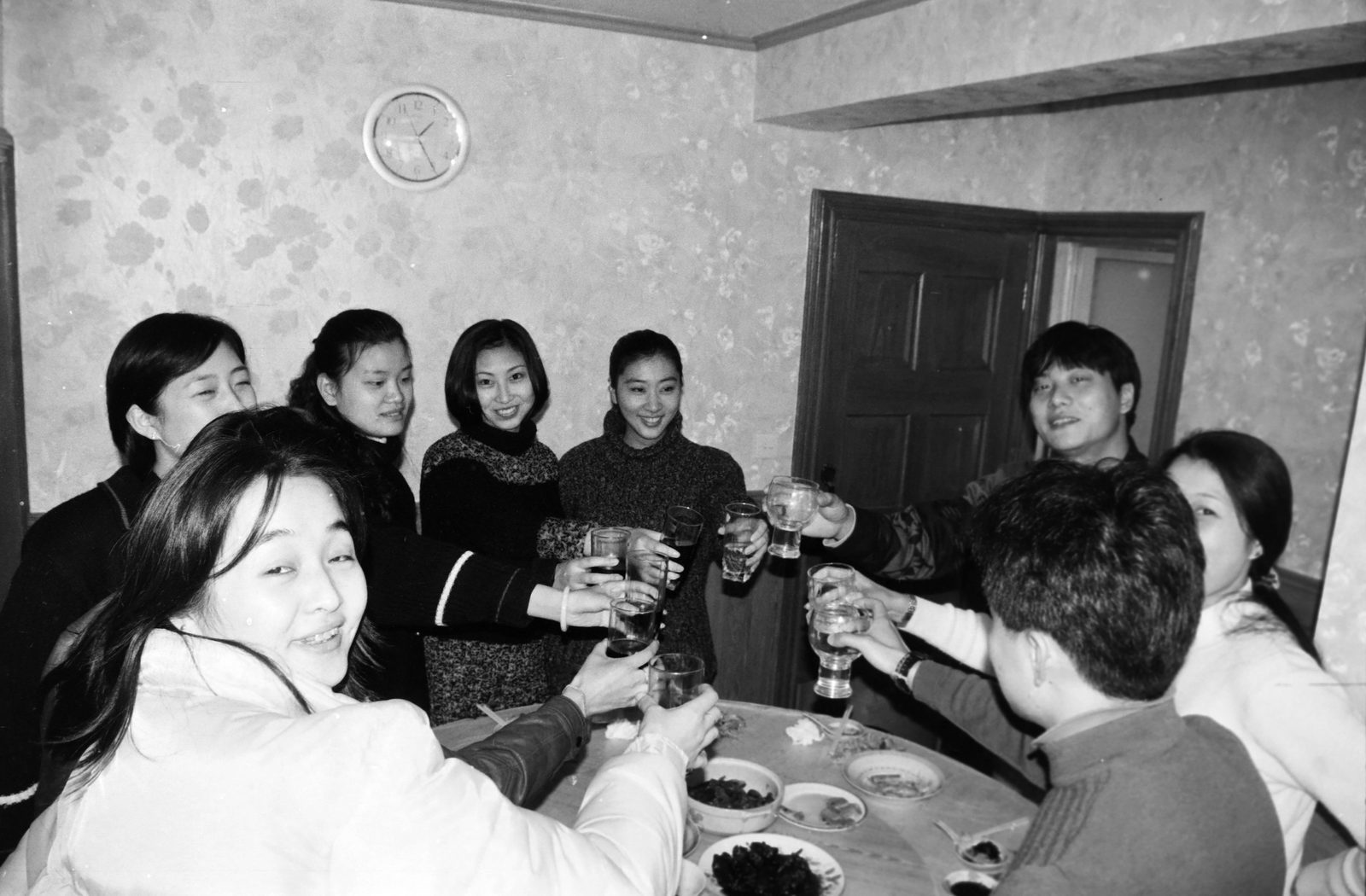 黑白照片：室内聚餐，八人围坐在圆桌旁举杯，墙上挂着一只时钟