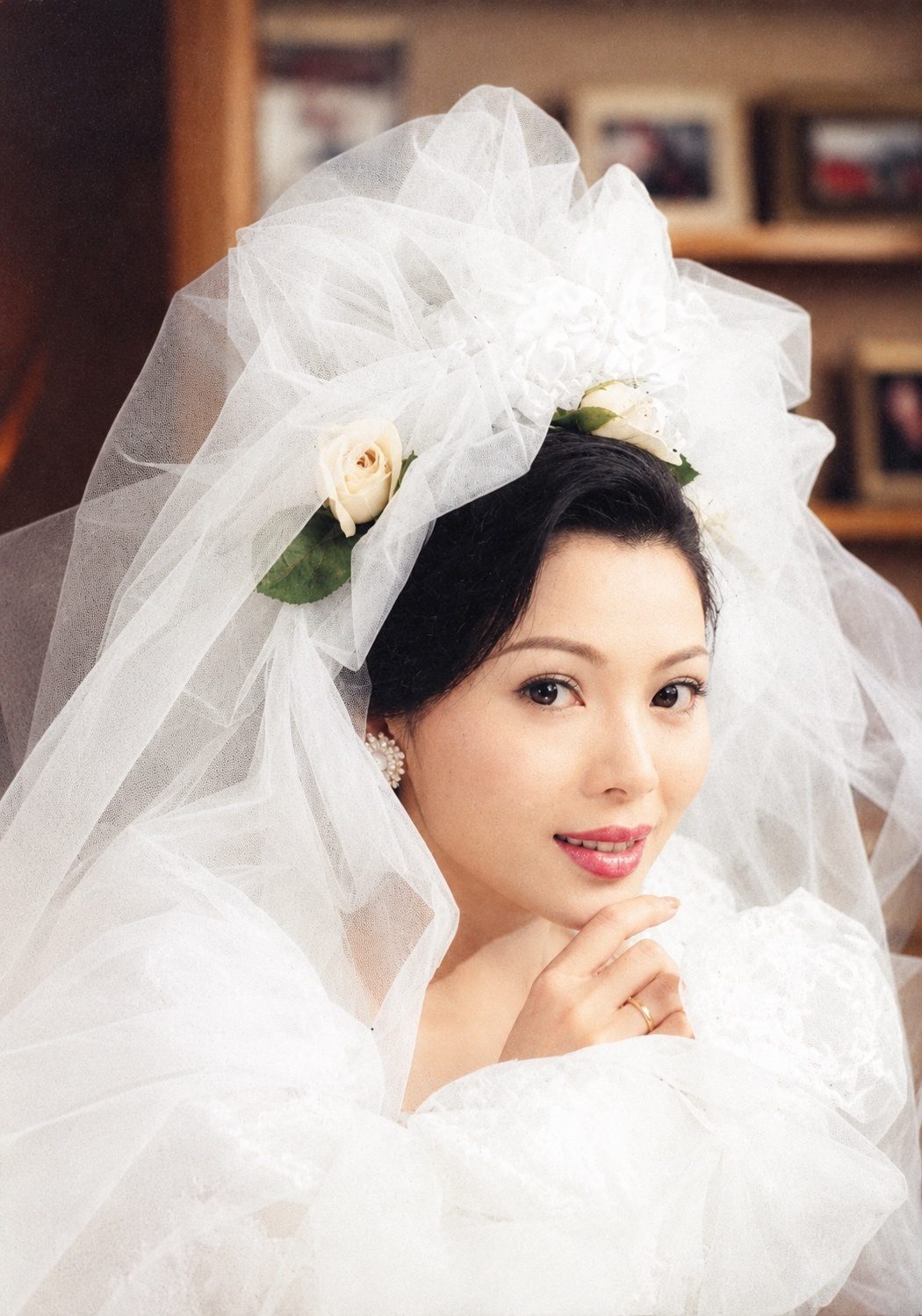 彩色照片：头戴缀着白玫瑰的白色头纱、身穿白色婚纱的女子，手指轻托下巴，微笑望向镜头