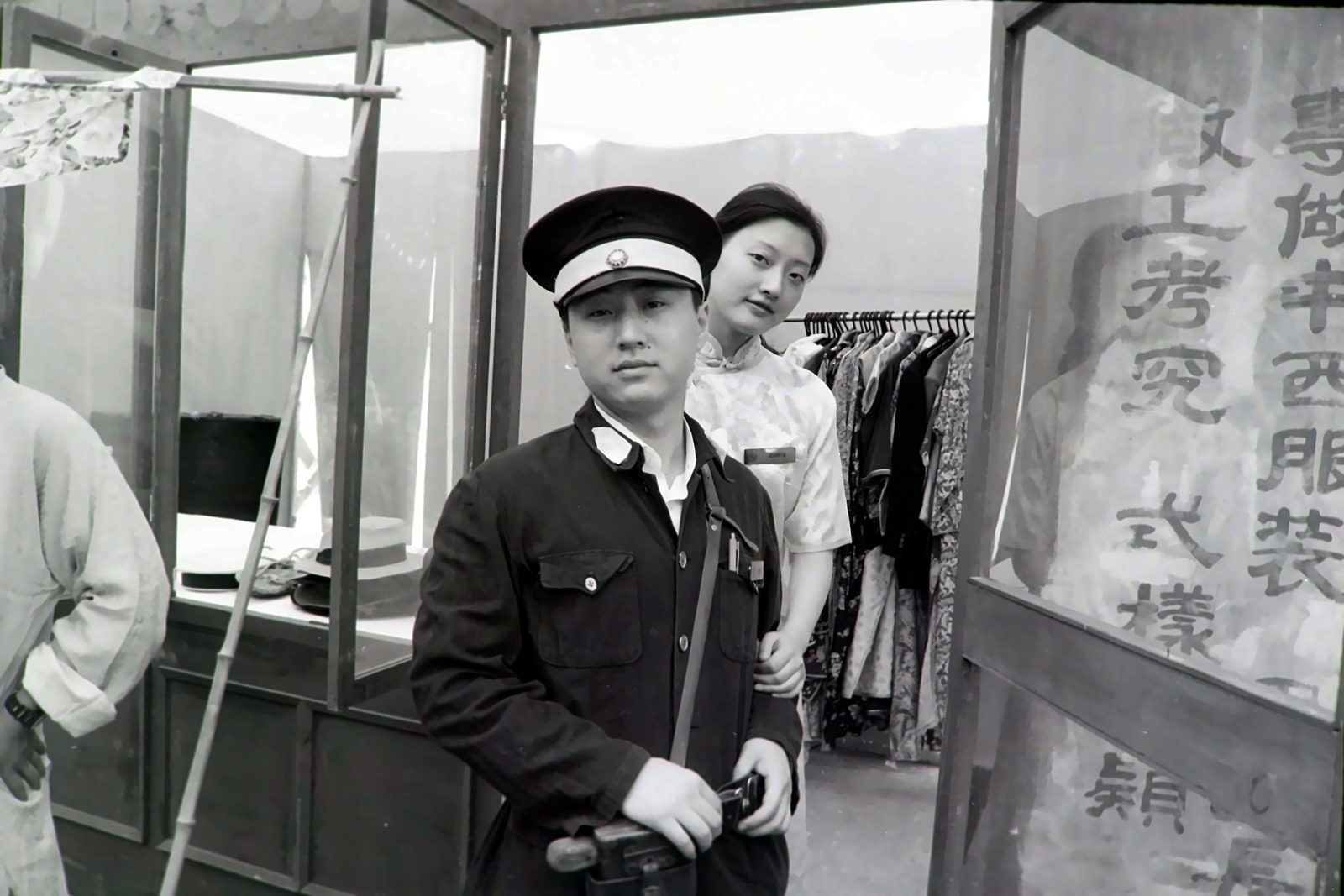 黑白照片：穿制服戴大檐帽的男子站在服装店门前，身后穿旗袍的女子探身望向镜头，右侧玻璃上写着中西服装的字样